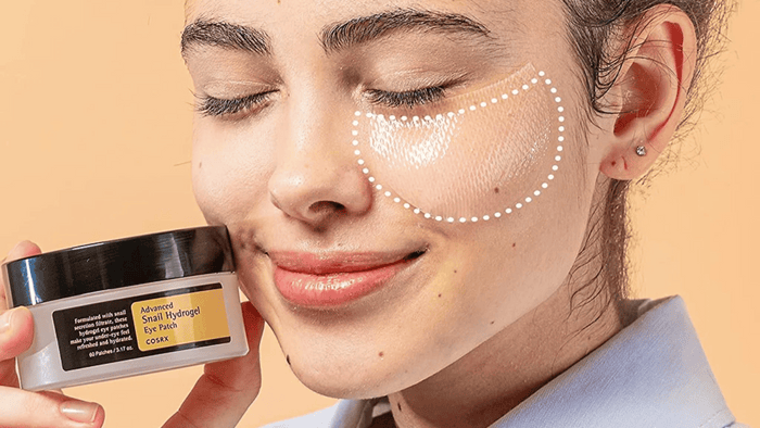 Kvinna som håller i en burk med eye patches från COSRX. Hon har även applicerat eye patches under högra ögat och ser ut att njuta av behandlingen.