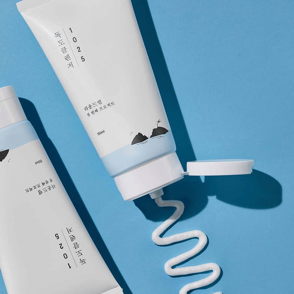 Dve tubi 1025 Dokdo Cleanser na modri podlagi, ena z odprtim pokrovčkom in ena z valovito linijo iztisnjenega čistila.