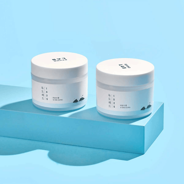 Dva bela kozarca 1025 Dokdo Pads, postavljena na svetlo modro polico, ki meče odsev, na modri podlagi.