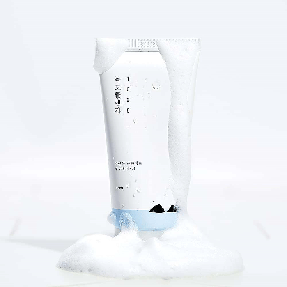 1025 Dokdo Cleanser v beli tubi, prekriti s peno, z vodnimi kapljicami in peno okoli dna, na svetli podlagi.