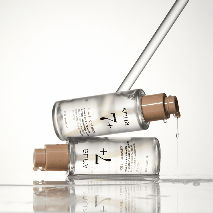 Slika prikazuje dve prozorni steklenički "Anua 77 Rice + Ceramide Hydrating Barrier Serum" s kapljicami, ki tečejo iz pipete. Serum, ki je obogaten z rižem in ceramidi, je namenjen vlaženju in krepitvi kožne pregrade. Stekleničke so elegantno oblikovane, kar poudarja občutek prestiža izdelka.