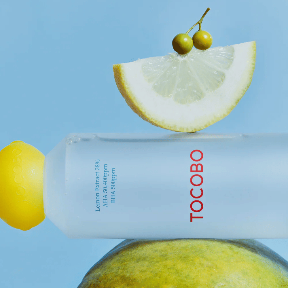 Steklenica TOCOBO AHA BHA Lemon Toner, okrašena s pokrovčkom v obliki limone. Rezina limone in majhne zelene jagode se uravnoteženo nahajajo na vrhu steklenice na modrem ozadju.