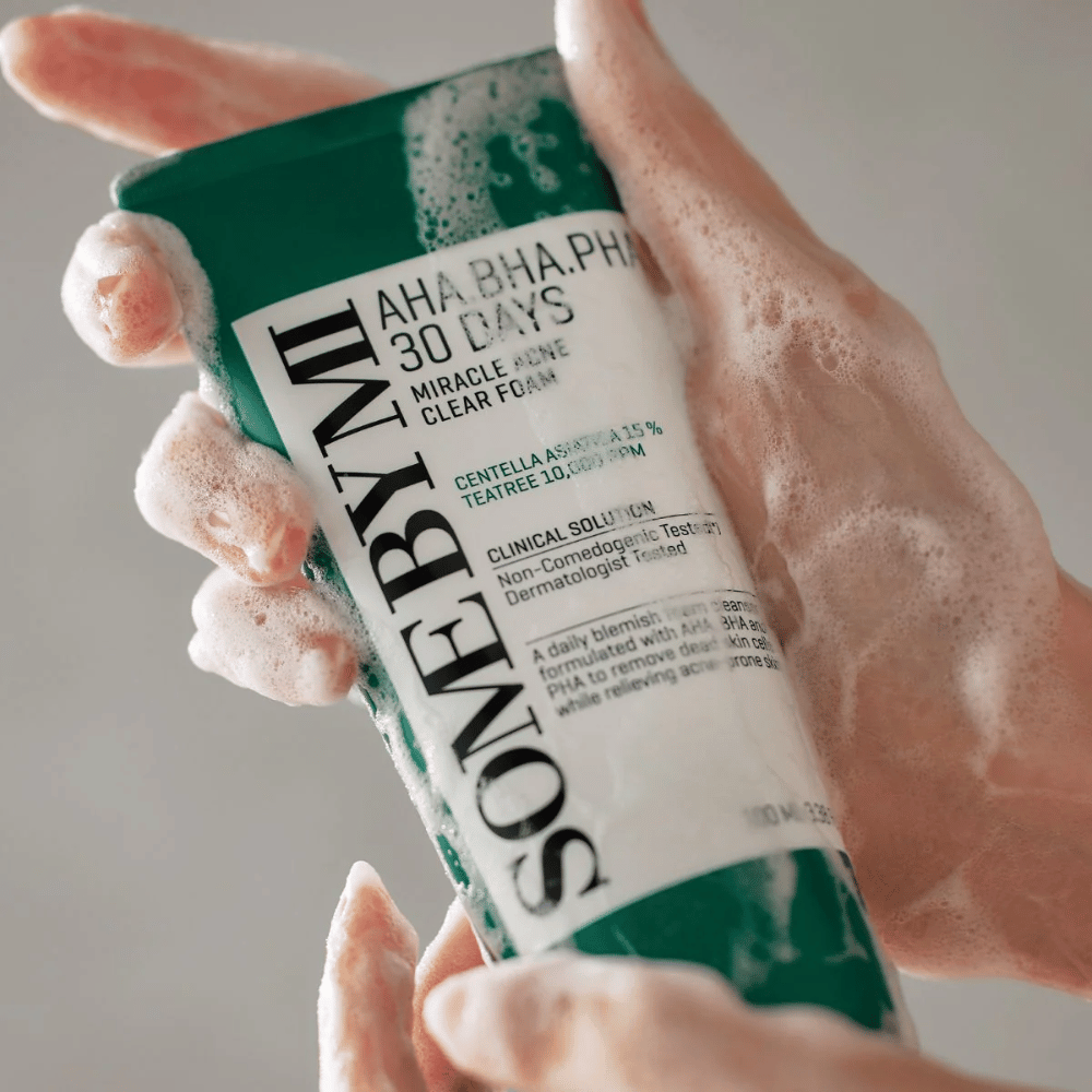 Oseba drži tubo "SOME BY MI AHA, BHA, PHA 30 Days Miracle Acne Clear Foam" s penastim vsebino nad rokami. Izdelek je namenjen za dnevno uporabo za boj proti aknam in čiščenje por s svojimi aktivnimi sestavinami kot so centella, olje čajevca in kisline kot AHA, BHA in PHA.