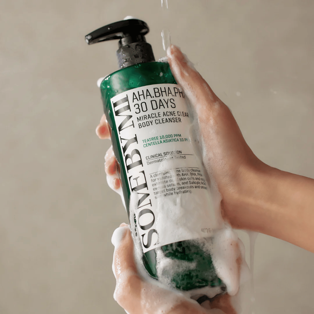 Oseba drži steklenico SOME BY MI AHA.BHA.PHA 30 Days Miracle Acne Clear Body Cleanser. Steklenica je zelena z belo etiketo in je prekrita s peno čistilnega sredstva. Oseba se umiva z izdelkom, medtem ko voda teče po rokah.