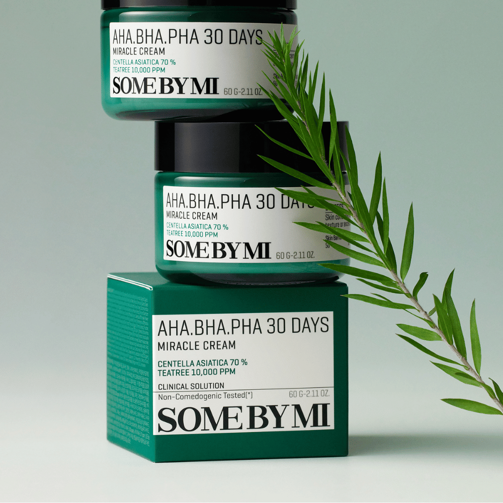 Slika, ki prikazuje dva kozarca "SOME BY MI AHA, BHA, PHA 30 Days Miracle Cream", naložena na zeleno obarvano škatlo. Krema, bogata s centella asiatica in čajevcem, je zasnovana za izboljšanje kakovosti kože in lajšanje težav s kožo. Deluje vlažilno in pomirjujoče.