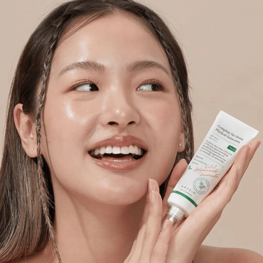 Slika prikazuje mlado žensko, ki se veselo smehlja in drži tubo AXIS-Y Complete No-Stress Physical Sunscreen. Ima sijoč in naraven videz z minimalnim ličenjem, kar poudarja izdelek, ki ga predstavlja.