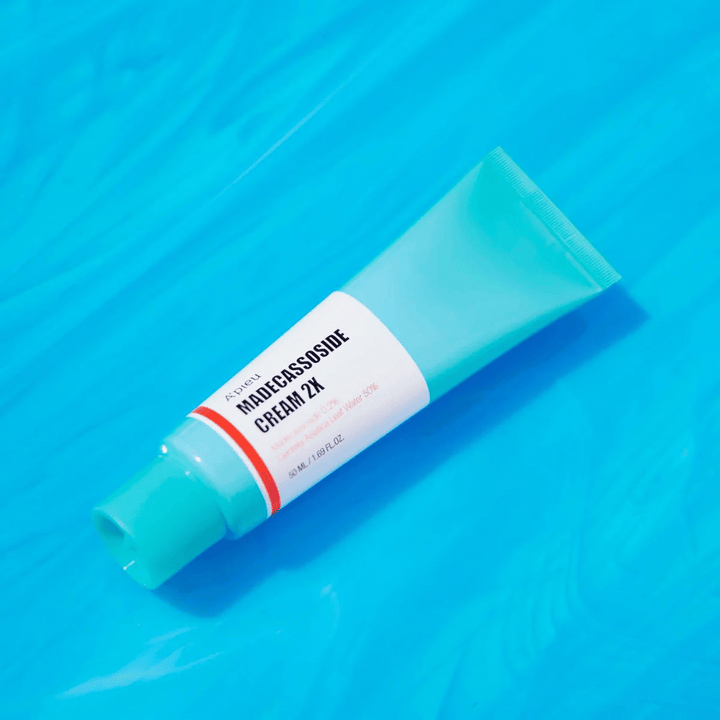 Tuba A'PIEU Madecassoside Cream 2X leži diagonalno čez modro ozadje z gubami, podobnimi vodi. Tuba ima belo telo z svetlo turkiznim pokrovčkom in rdeče do oranžno obarvano etiketo, kjer sta ime izdelka in aktivna sestavina Madecassoside 2% jasno napisana. Na etiketi je tudi navedena količina, ki je 50 ml / 1.8 fl oz. Svetla in barvita predstavitev daje svež in energičen občutek.