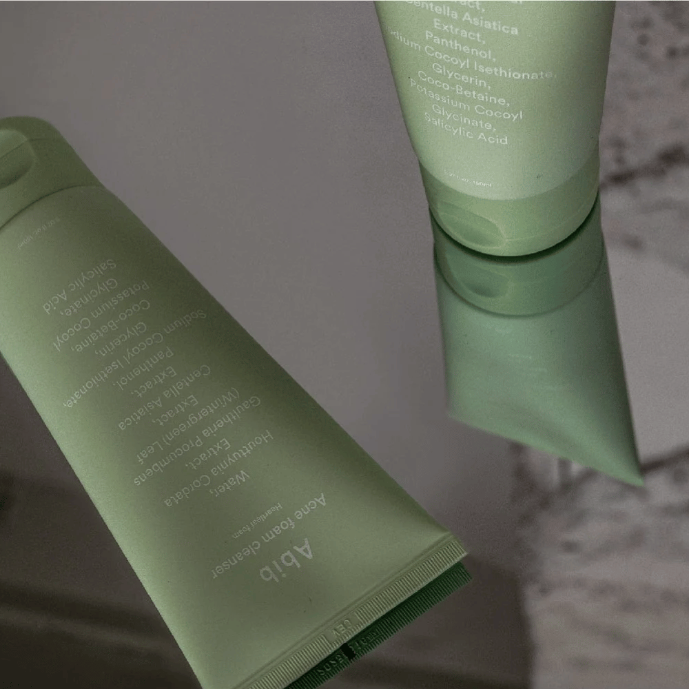 Zelena tuba Abib Acne Foam Cleanser Heartleaf, ležeča na zrcalni mizi s sprednjo stranjo izdelka in odsevom besedila. Sestavine, kot so salicilna kislina, centella in ekstrakt srčastega lista, so jasno vidne na embalaži.