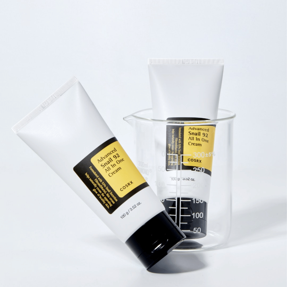 Tuba COSRX Advanced Snail 92 All in One Cream leži poleg obrnjene prozorne merilne cilindre na beli podlagi.