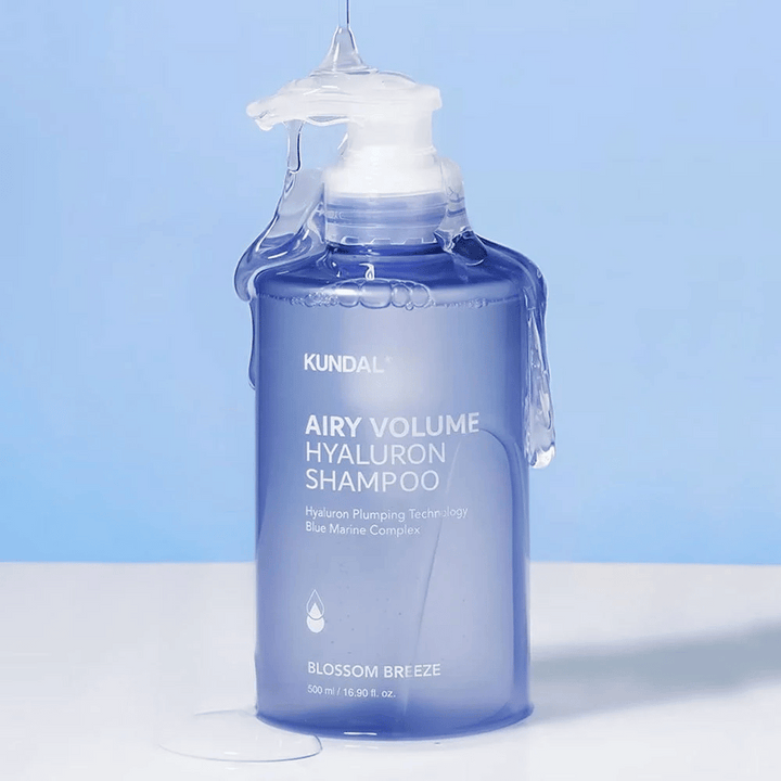 KUNDAL Airy Volume Hyaluron Shampoo Blossom Breeze v modri steklenički s črpalko, s hialuronom in Blue Marine Complex, fotografirano s tekočo vodo.