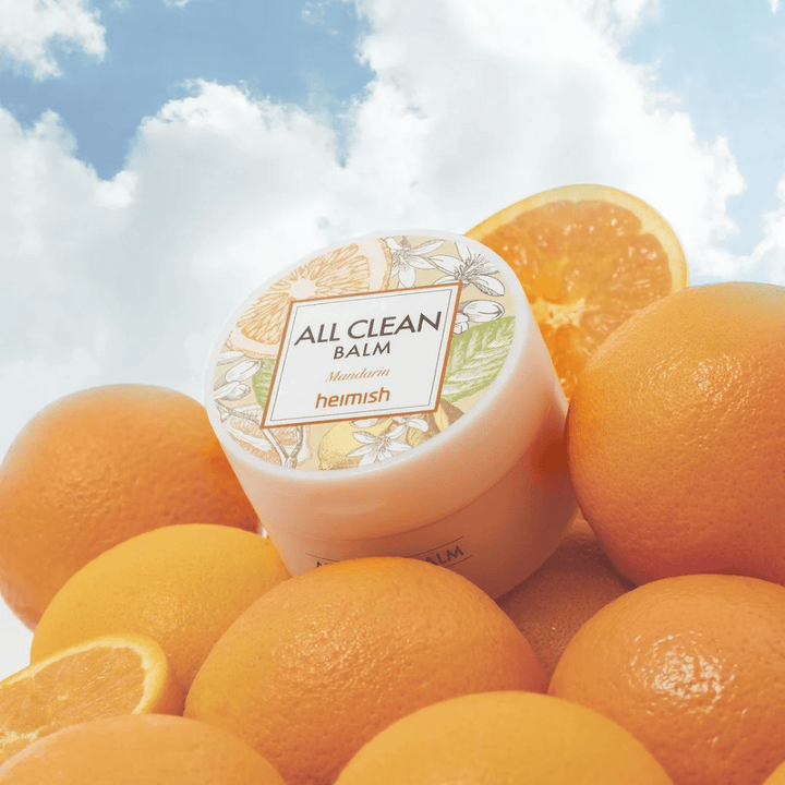 Kozarec Heimish All Clean Balm Mandarin postavljen na kup pomaranč, z modrim nebom v ozadju.