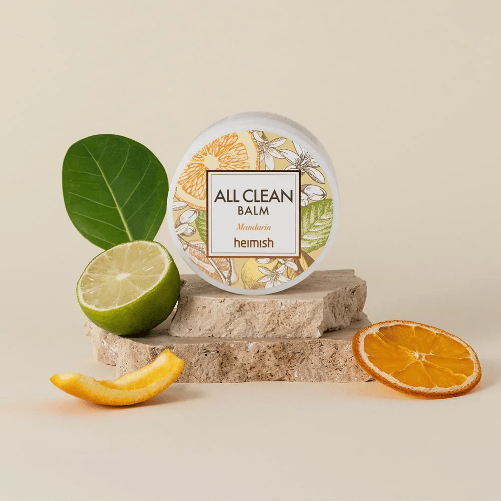 Kozarec Heimish All Clean Balm Mandarin postavljen na kamne, obdan z mandarinino lupino in citrusovimi listi, pred svetlim ozadjem.