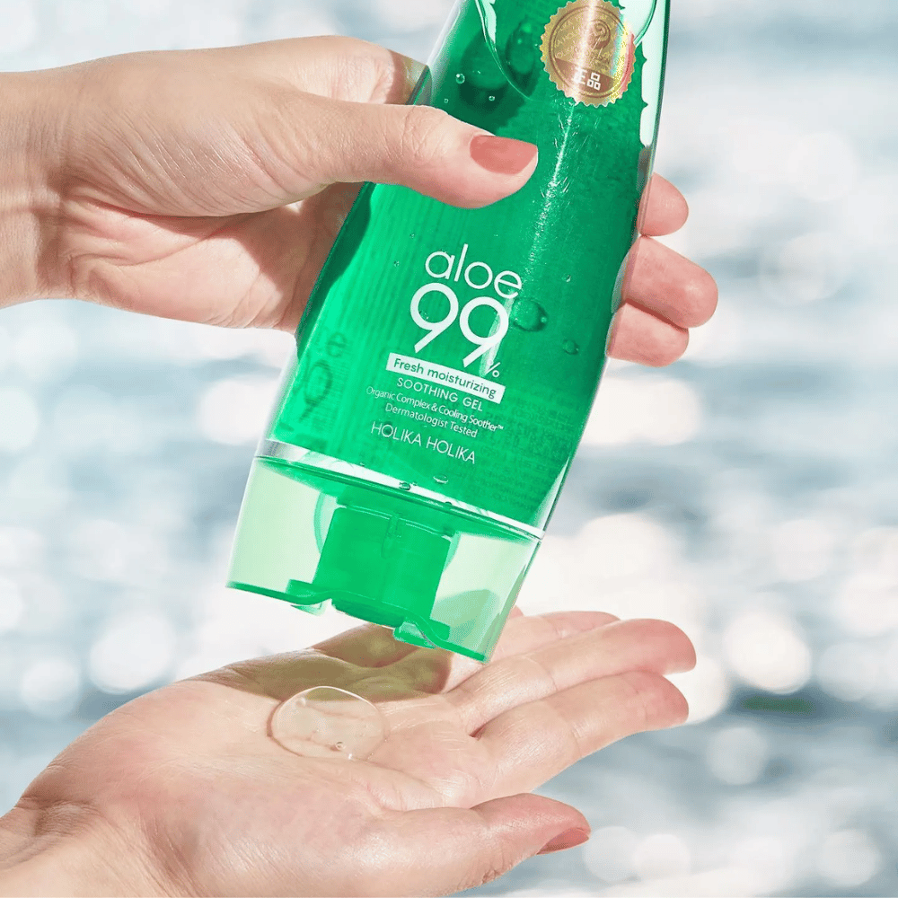 Roka drži zeleno tubo z Holika Holika Aloe 99% Soothing Gel, medtem ko druga roka ujame klik gela, ki se iztisne. Ozadje je zamegljeno in prikazuje vodni, bleščeči vzorec.
