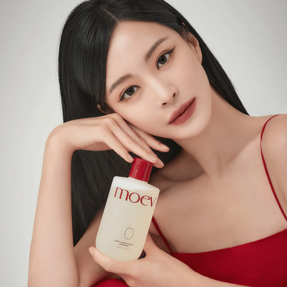 Ženska z dolgimi, sijočimi lasmi drži MOEV Annurcatin Shampoo – korejski šampon, ki vlaži, krepi in ščiti lase z naravnimi sestavinami.