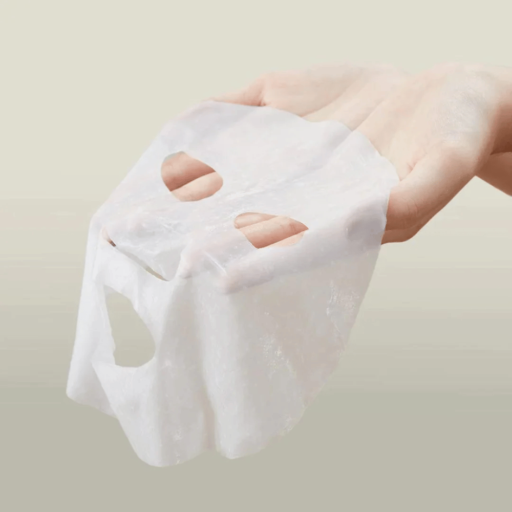 Slika prikazuje roko, ki drži obrazno masko znamke "Anua". Zdi se, da gre za "Heartleaf Cream Mask Night Solution", kar nakazuje, da je to nočna maska za nego kože z izvlečkom srčastega lista. Maska je videti iz blaga in je prepojena s kremo ali serumom, kar je značilno za korejske izdelke za nego kože, ki se osredotočajo na intenzivno vlaženje in nego kože ponoči.