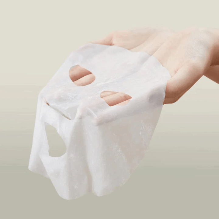 Slika prikazuje roko, ki drži obrazno masko znamke "Anua". Zdi se, da gre za "Heartleaf Cream Mask Night Solution", kar nakazuje, da je to nočna maska za nego kože z izvlečkom srčastega lista. Maska je videti iz blaga in je prepojena s kremo ali serumom, kar je značilno za korejske izdelke za nego kože, ki se osredotočajo na intenzivno vlaženje in nego kože ponoči.