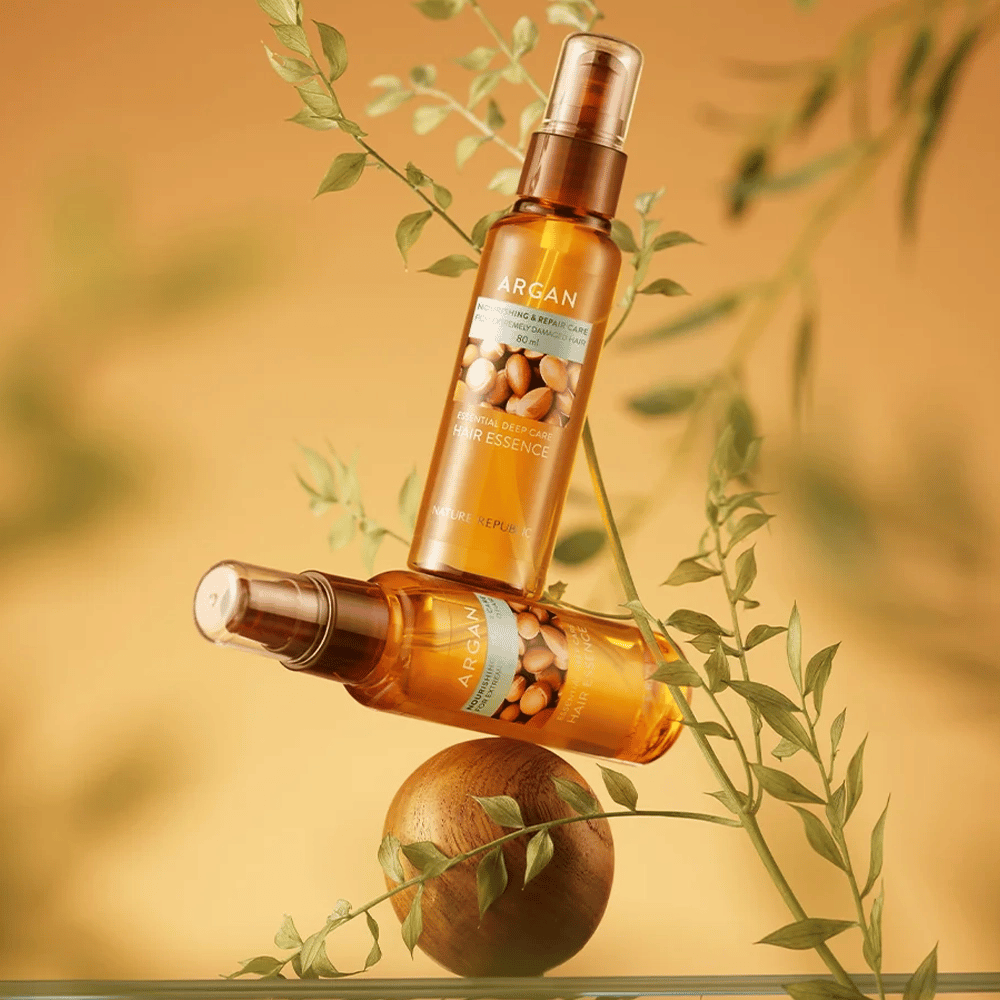 Dve steklenički Nature Republic Argan Essential Moisture Hair Oil postavljeni med zelenimi listi – svilnat sijaj za suhe lase.