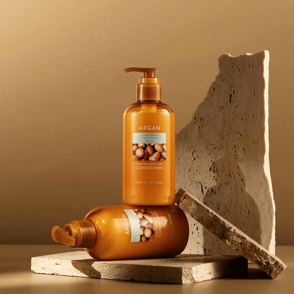 Dve steklenici Nature Republic Argan Essential Moisture Repairing Conditioner na kamniti podlagi v toplem tonu.