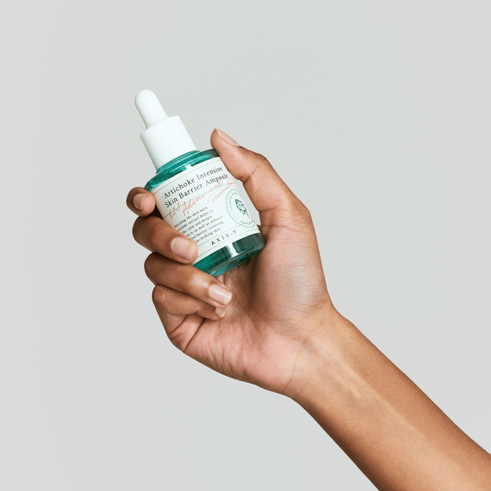 Roka drži steklenico seruma za nego kože z etiketo "Artichoke Intensive Skin Barrier Ampoule."