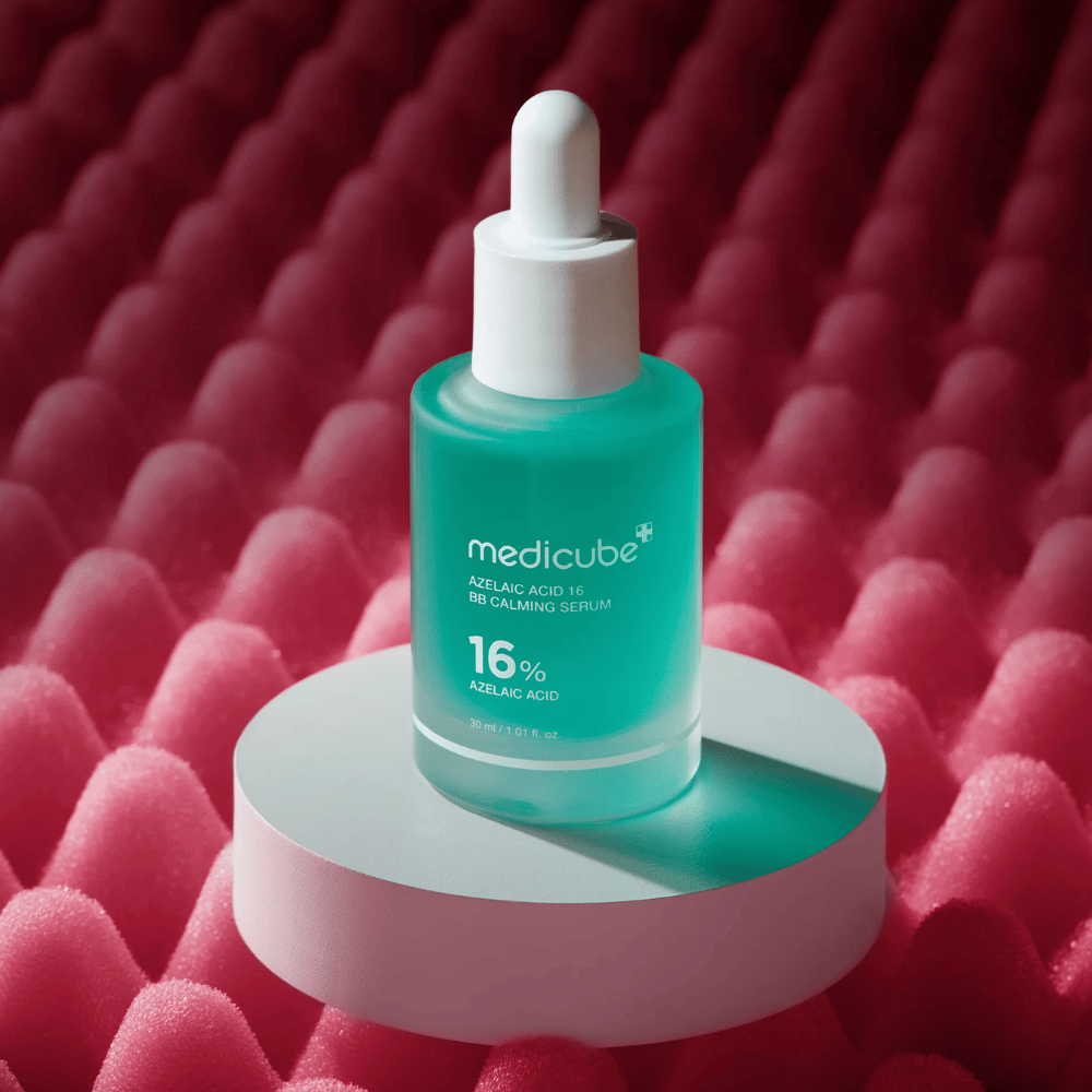 Slika izdelka Medicube Azelaic Acid 16 BB Calming Serum postavljena na belo podijsko površino z rožnatim strukturiranim ozadjem, ki poudarja pomirjujoč in kožo izboljšujoč profil seruma.