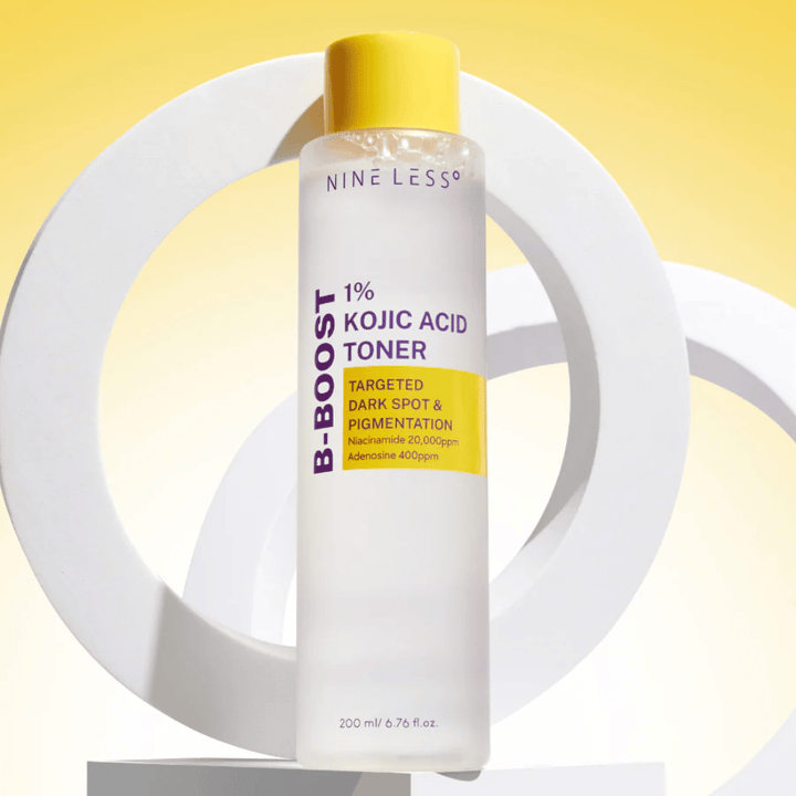 NINE LESS B-Boost 1% Kojic Acid Toner postavljen pred rumeno ozadje z besedilom "Targeted Dark Spot & Pigmentation". Tonik za izenačevanje tena kože z niacinamidom in adenozinom.