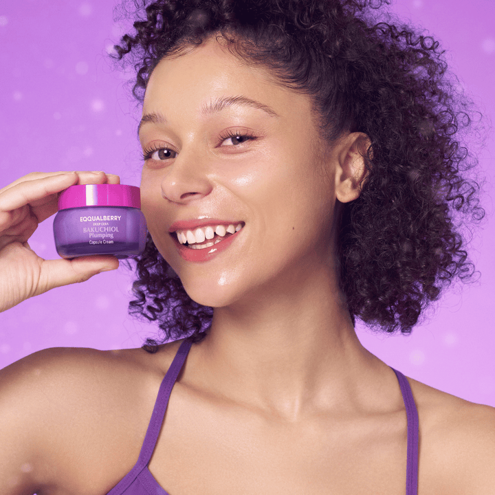 Ženska z žarečo kožo drži EQQUALBERRY Bakuchiol Plumping Capsule Cream in se smehlja pred vijoličnim ozadjem.