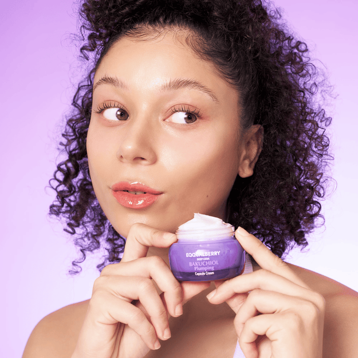 Ženska drži EQQUALBERRY Bakuchiol Plumping Capsule Cream v roki in razkazuje svojo navlaženo, sijočo kožo proti vijoličnemu ozadju.