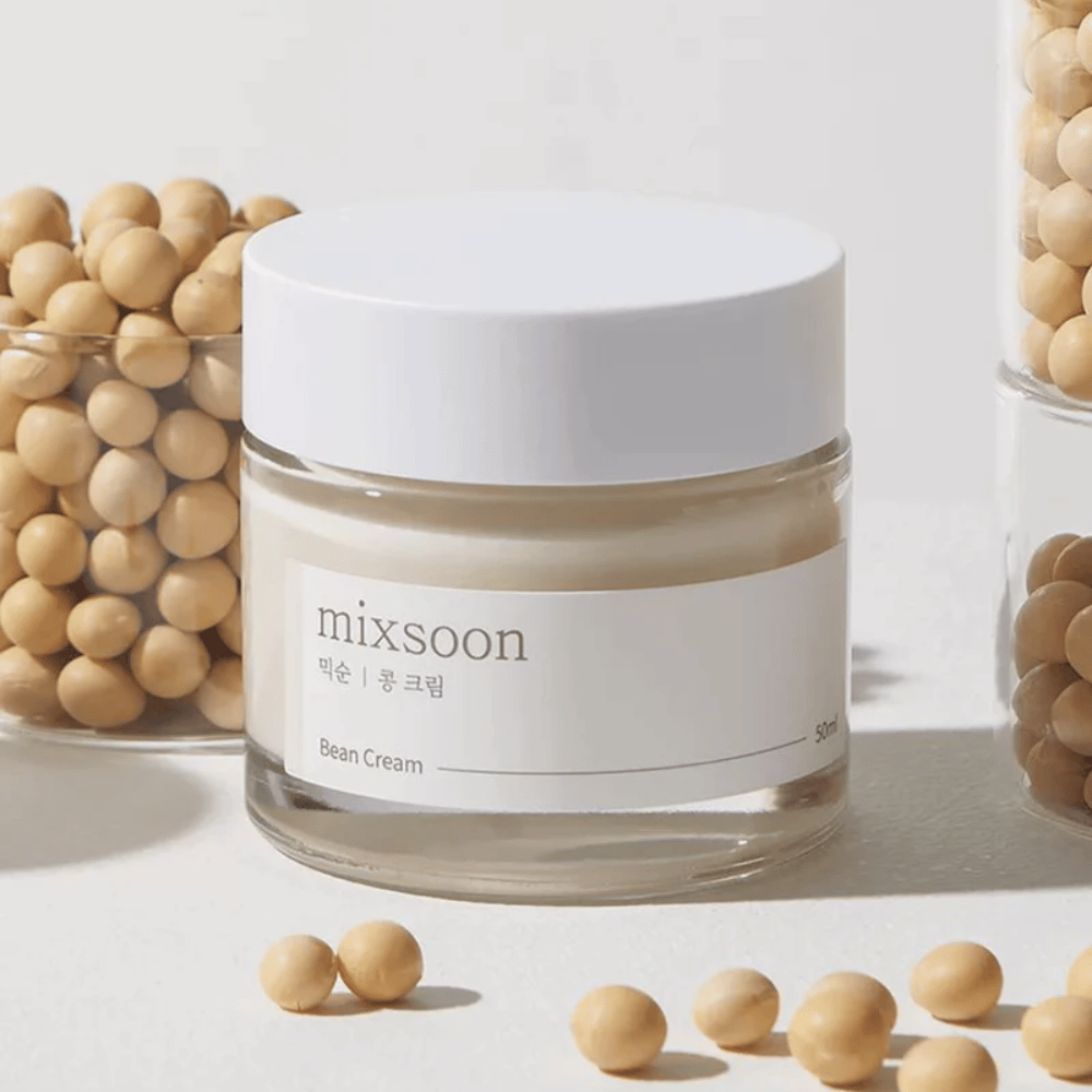 Prozoren kozarec z belo etiketo in pokrovom, označen z "mixsoon 팥 크림" in "Bean Cream" z navedeno prostornino "50ml". Kozarec vsebuje svetlo rumeno kremo in je postavljen na svetlo površino. Poleg kozarca je steklena posoda polna svetlo rjavih fižolov, ki so raztreseni po površini, kar nakazuje, da krema vsebuje fižolov ekstrakt. Ozadje in osvetlitev ustvarjata toplo in naravno vzdušje.