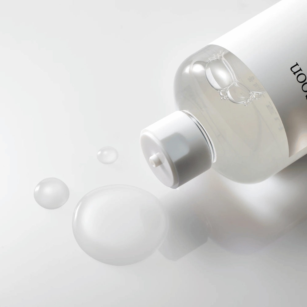 Prozorna steklenica leži na boku na sijoči površini z lahkim tonerjem, ki počasi teče ven in tvori kapljice ter večji mehurček na površini. Steklenica ima belo etiketo in bel črpalni mehanizem. Osvetlitev je mehka in ustvarja čist in pomirjujoč ambient, medtem ko je tekoča konsistenca tonika in njegova jasnost poudarjena s tem, kako se širi po površini.