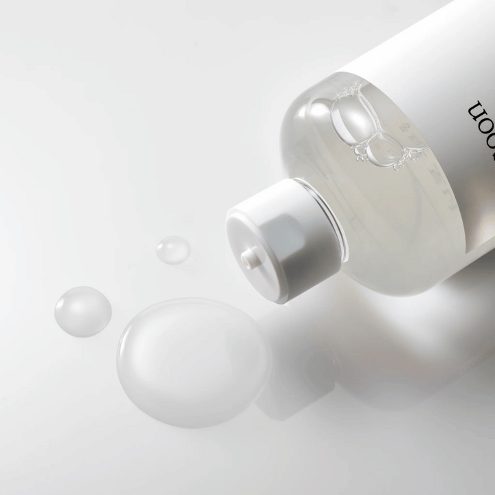 Prozorna steklenica leži na boku na sijoči površini z lahkim tonerjem, ki počasi teče ven in tvori kapljice ter večji mehurček na površini. Steklenica ima belo etiketo in bel črpalni mehanizem. Osvetlitev je mehka in ustvarja čist in pomirjujoč ambient, medtem ko je tekoča konsistenca tonika in njegova jasnost poudarjena s tem, kako se širi po površini.