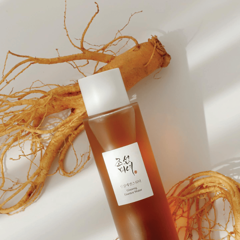 Rjava steklenica Beauty of Joseon Ginseng Essence Water z belim pokrovčkom leži poleg sveže korenine ginsenga na beli podlagi.