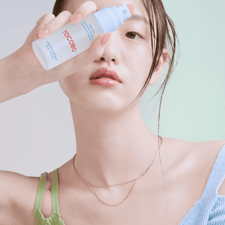 Mlada ženska z nežnim ličenjem in naravnim tenom kože drži stekleničko TOCOBO Bifida Biome Essence pred obrazom. Oblečena je v svetlo modro majico z zelenimi naramnicami in tanko zlato verižico proti svetlemu ozadju.