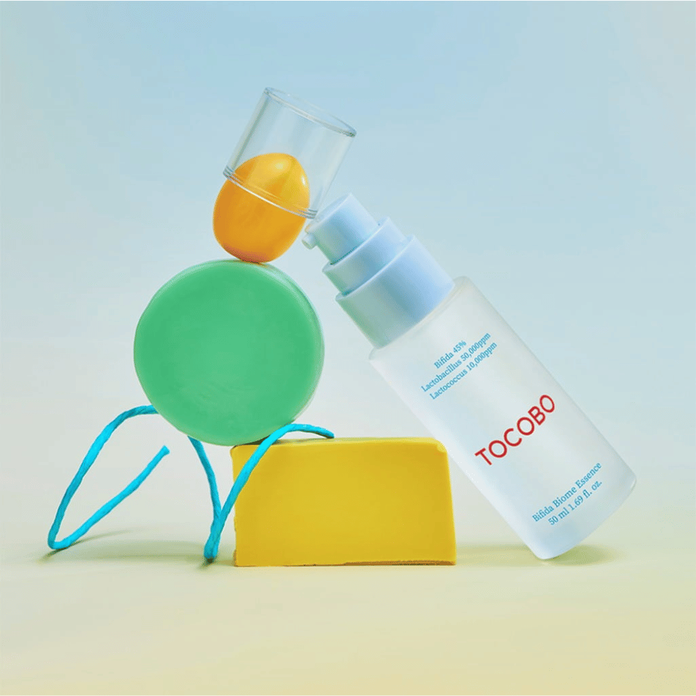 TOCOBO Bifida Biome Essence v zmrznjeni steklenički s črpalko z rdečim logotipom, postavljena na barvito ozadje z zelenimi, rumenimi in oranžnimi geometrijskimi oblikami. Besedilo na steklenički navaja Bifida 5 %, Lactobacillus 3 000 ppm in Lactococcus 1 000 ppm.
