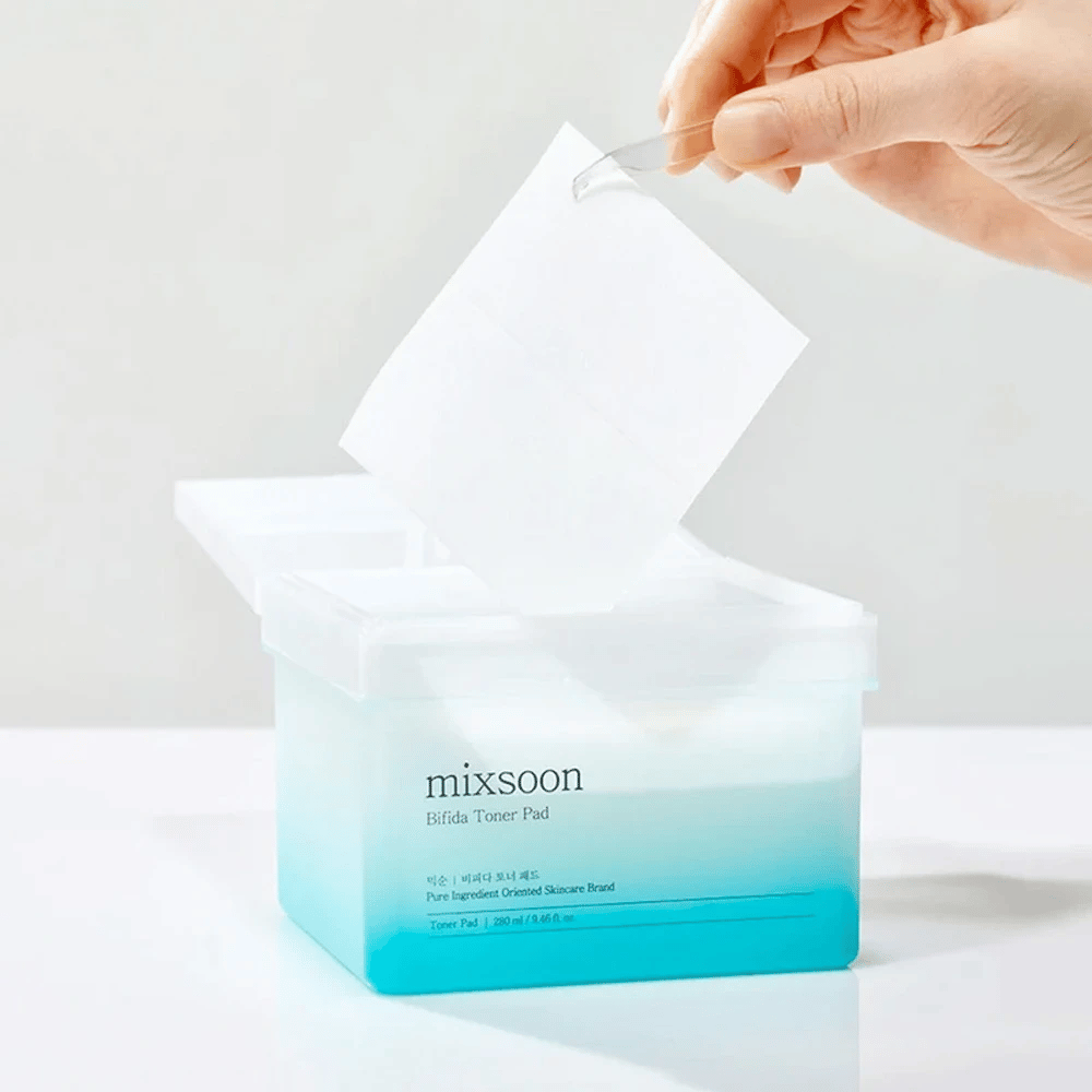 Prozorna posoda z modro in belo gradiento, ki vsebuje Bifida tonik blazinice. Roka vzame tonik blazinico iz posode, ki je označena z "mixsoon Bifida Toner Pad" in vsebuje 280 ml (9,46 fl. oz.) tonik blazinic, namenjenih za nego kože.