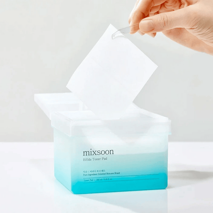 Prozorna posoda z modro in belo gradiento, ki vsebuje Bifida tonik blazinice. Roka vzame tonik blazinico iz posode, ki je označena z "mixsoon Bifida Toner Pad" in vsebuje 280 ml (9,46 fl. oz.) tonik blazinic, namenjenih za nego kože.