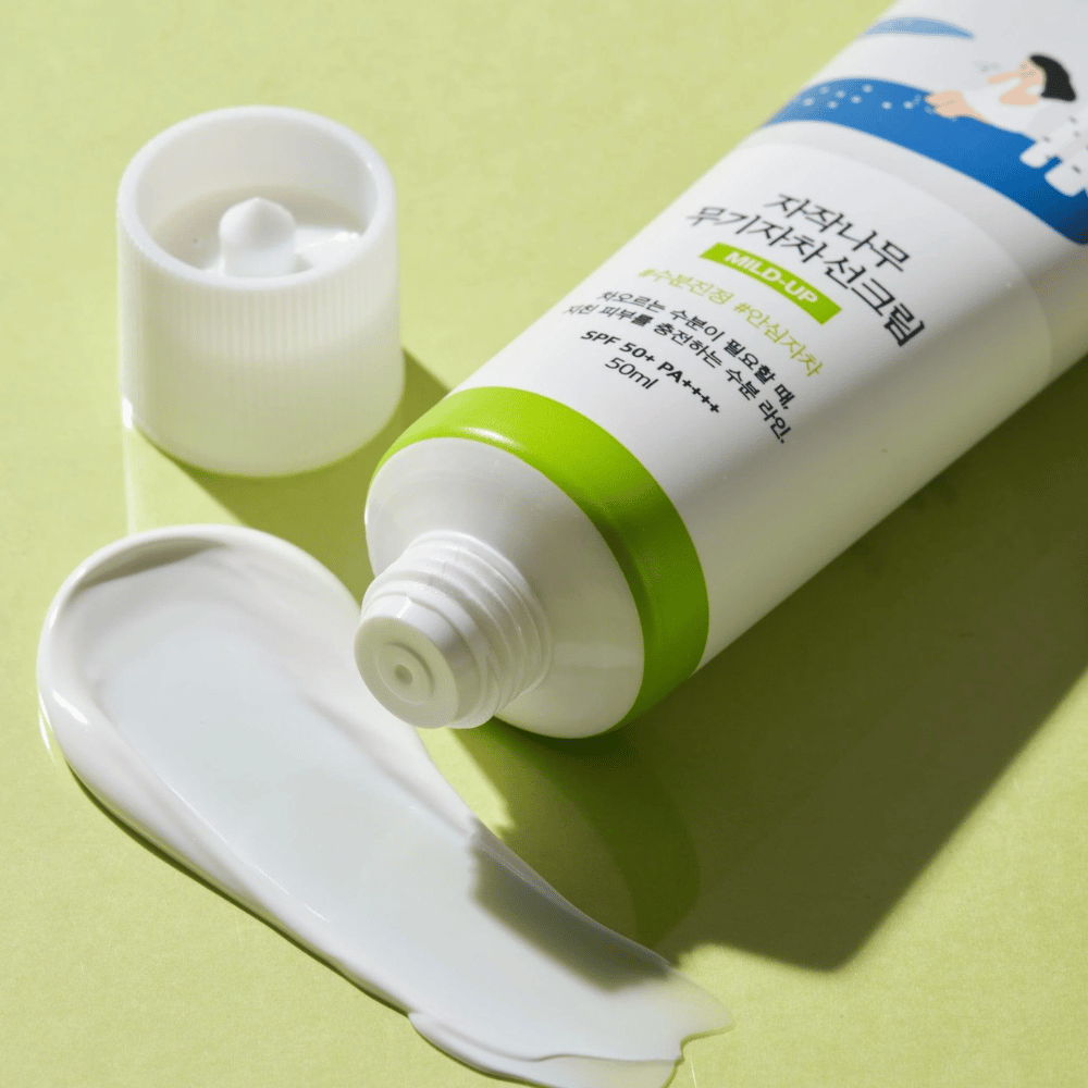 Pogled od blizu na Birch Juice Moisturizing Mild Up Sunscreen z odvijačenim pokrovčkom, bela sončna krema razmazana na svetlo zeleni podlagi, SPF 50+ PA++++, 50 ml.
