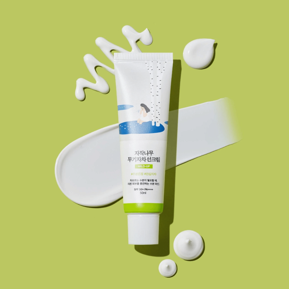 Birch Juice Moisturizing Mild Up Sunscreen – bela tuba z zeleno osnovo na zeleni podlagi, obdana z razmazano sončno kremo in kapljicami, SPF 50+ PA++++, 50 ml.