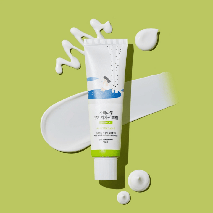 Birch Juice Moisturizing Mild Up Sunscreen – bela tuba z zeleno osnovo na zeleni podlagi, obdana z razmazano sončno kremo in kapljicami, SPF 50+ PA++++, 50 ml.