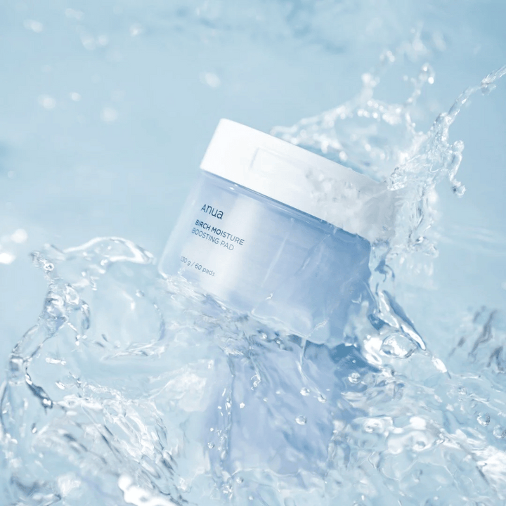 Slika Anuas "Birch Moisture Boosting Pad" v prozorni modri posodi s kapljicami vode okoli, kar nakazuje na vlažilni učinek izdelka.