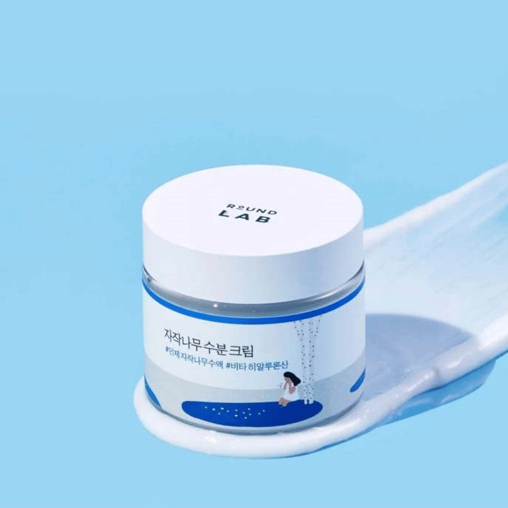 Round Lab Birch Juice Moisturizing Cream v modrikasti posodi z belim pokrovom, ki stoji v sloju kreme na modri podlagi.