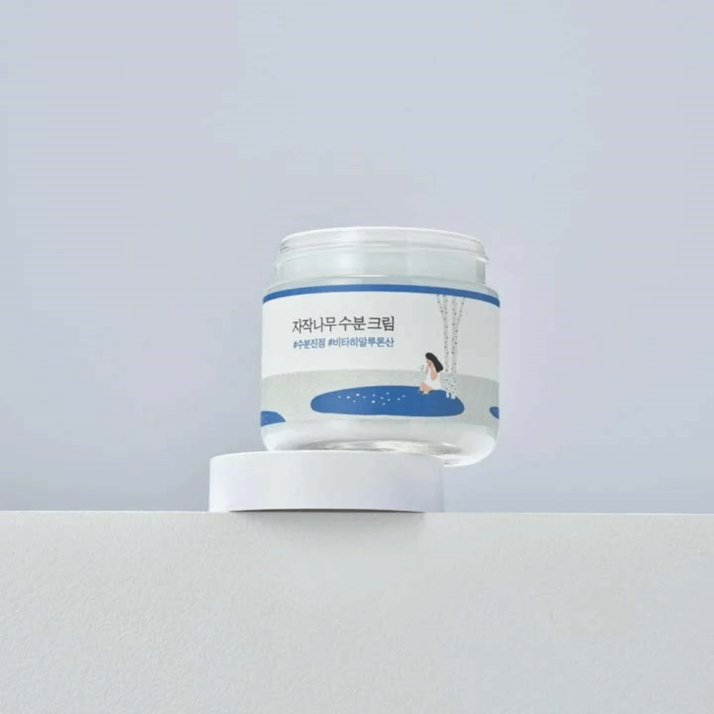 Round Lab Birch Juice Moisturizing Cream v prozorni posodi z belo etiketo in modro ilustracijo ženske pri brezi, ki stoji na beli površini proti svetli podlagi.