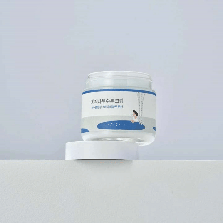 Round Lab Birch Juice Moisturizing Cream v prozorni posodi z belo etiketo in modro ilustracijo ženske pri brezi, ki stoji na beli površini proti svetli podlagi.