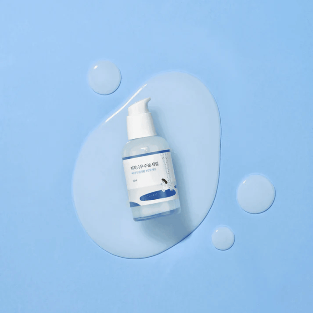 Round Lab Birch Juice Moisturizing Serum v prozorni steklenički s črpalko postavljen na modro ozadje s kapljicami in razlitim serumom.