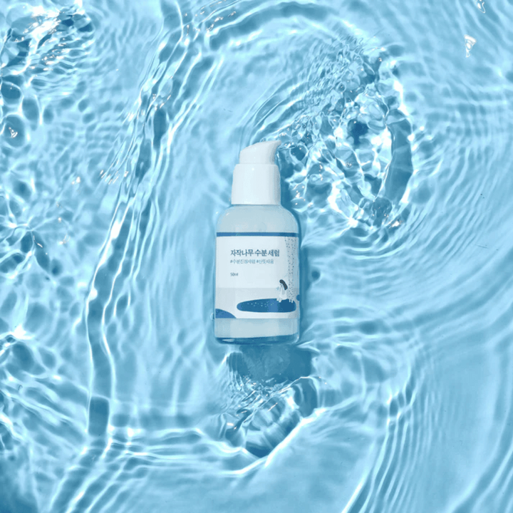 Round Lab Birch Juice Moisturizing Serum v steklenički s črpalko, ki plava na modri vodi z odsevi sončne svetlobe.