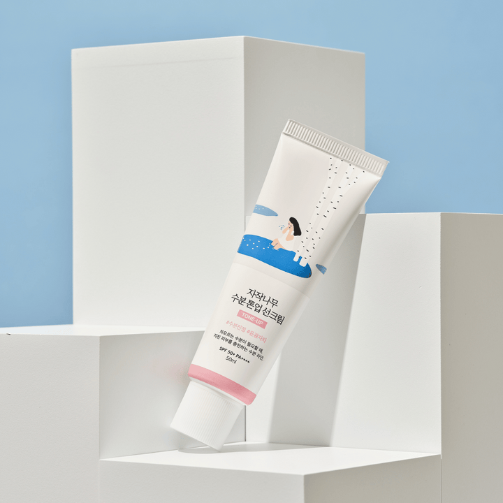 Tuba z Birch Juice Moisturizing Tone Up Sunscreen, postavljena na belih kockah proti svetlo modrem ozadju. Tuba je bela z ilustracijo ženske pri brezi in vodi. Besedilo v korejščini navaja, da ima izdelek SPF 50+ PA++++ in vsebuje 50 ml.