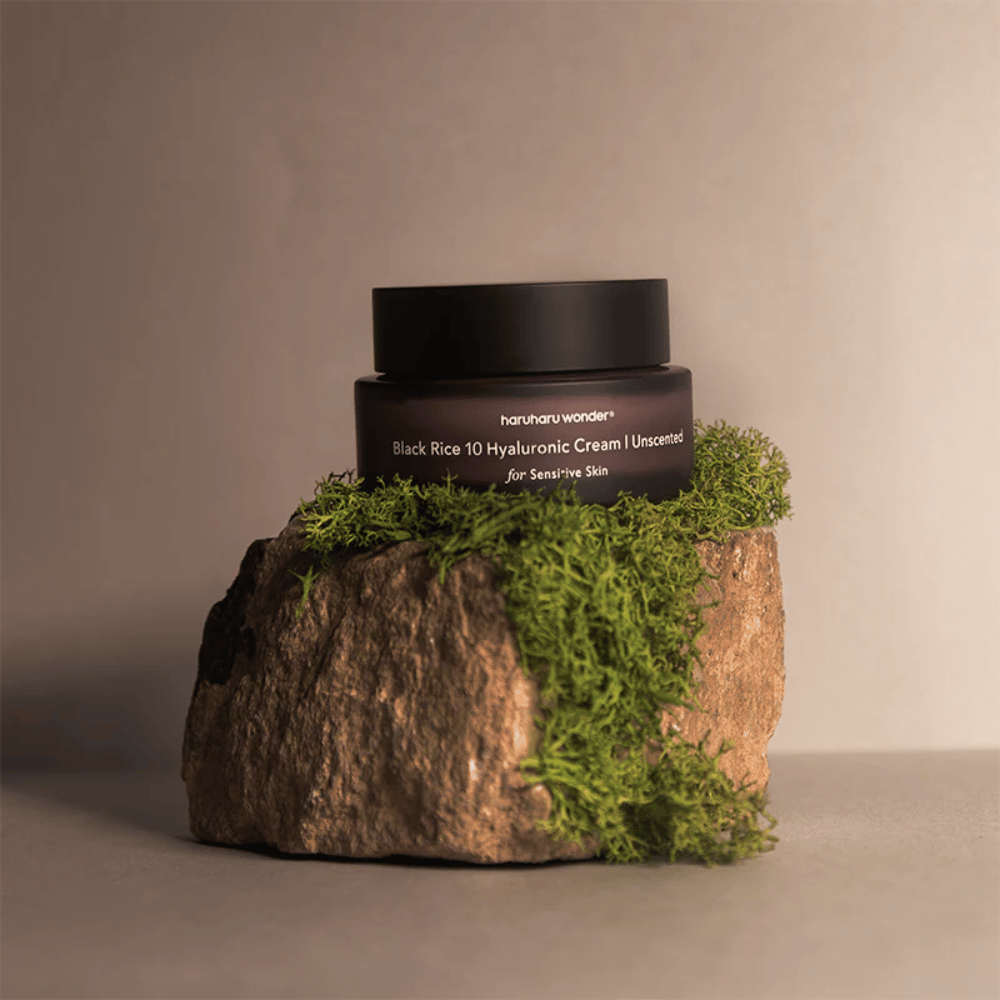 Slika prikazuje kozarec kreme za kožo z oznako "haruharu wonder Black Rice 10 Hyaluronic Cream Unscented for Sensitive Skin". Kozarec je črn in stoji na veliki skali, pokriti z svetlo zelenim mahom, kar ustvarja naraven in zemeljski občutek.