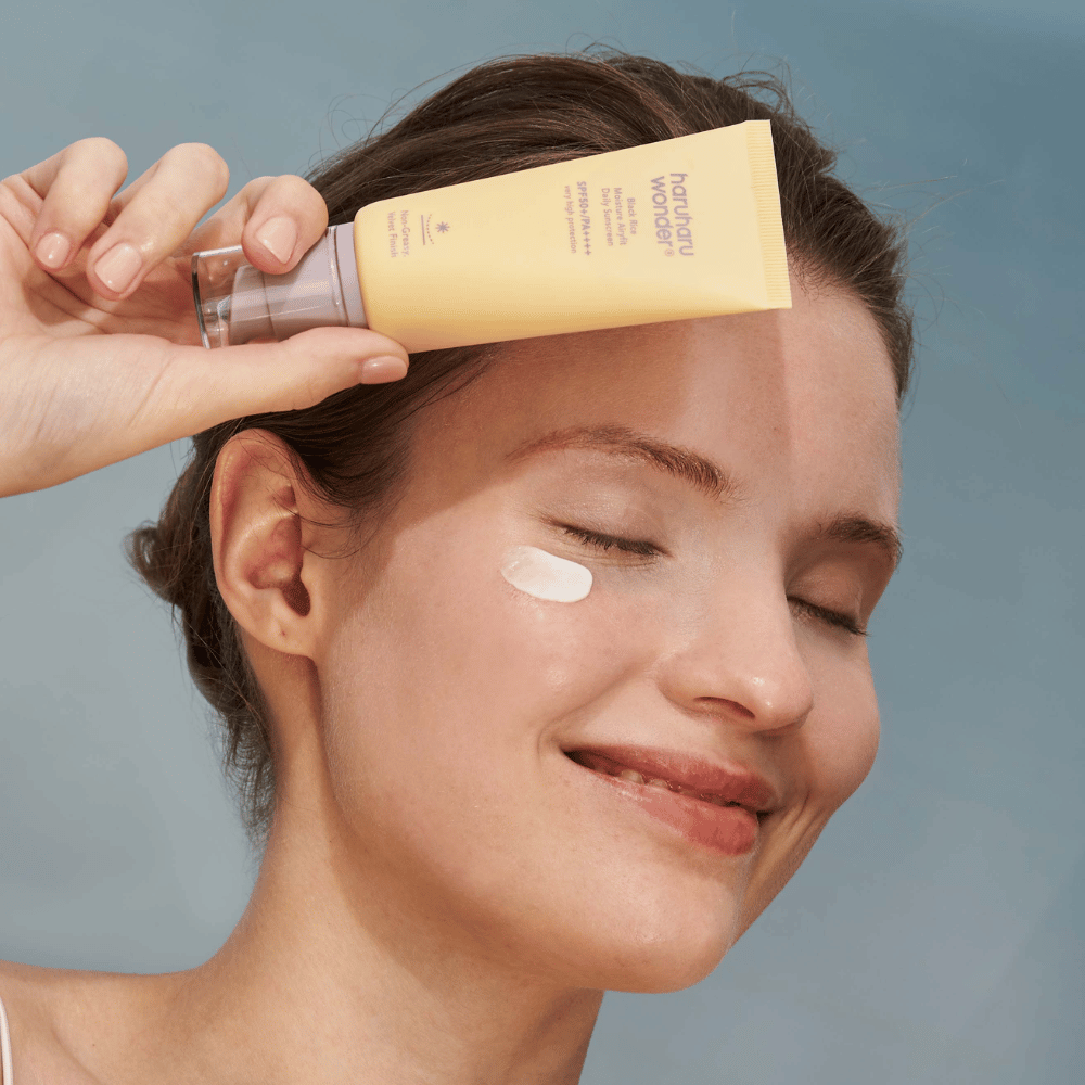 Nasmejana ženska nanese Black Rice Moisture Airyfit Daily Sunscreen SPF 50+ PA++++ iz Haruharu Wonder na svoj obraz. Drži rumeno tubo s kremo za sončenje in ima kapljico izdelka na licu. Ozadje je svetlo modro.
