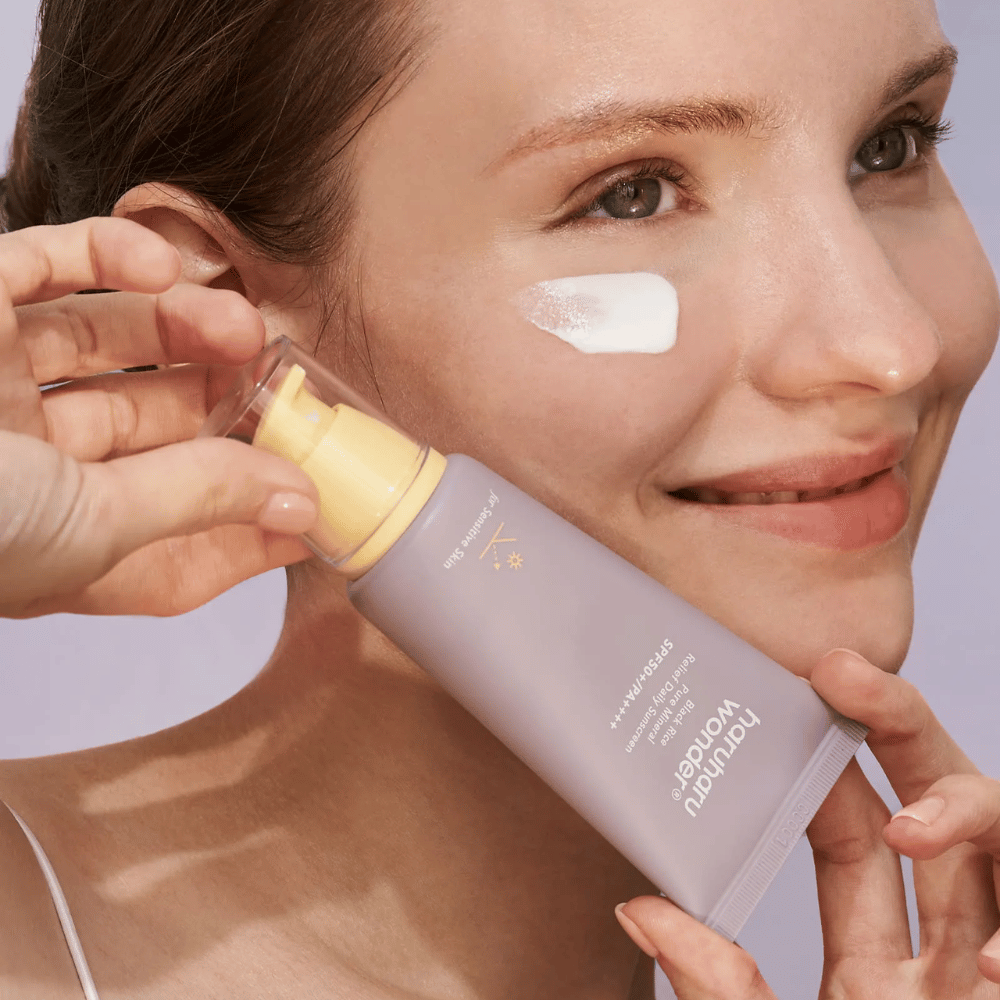 Nasmejana ženska nanese Haruharu Wonder Black Rice Pure Mineral Relief Daily Sunscreen SPF 50+ PA++++ na svoj obraz. Drži vijolično tubo s kremo za sončenje in ima kapljico izdelka na licu. Ozadje je svetlo vijolično.