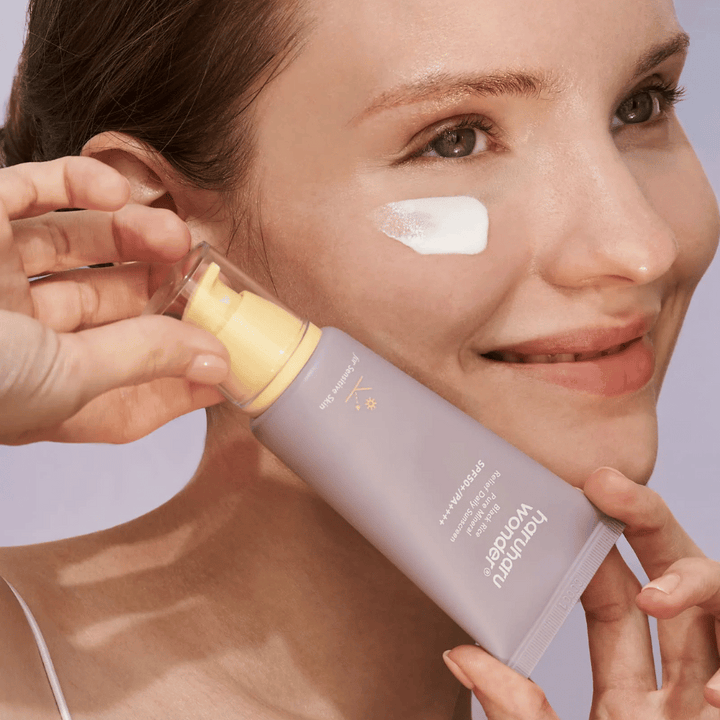 Nasmejana ženska nanese Haruharu Wonder Black Rice Pure Mineral Relief Daily Sunscreen SPF 50+ PA++++ na svoj obraz. Drži vijolično tubo s kremo za sončenje in ima kapljico izdelka na licu. Ozadje je svetlo vijolično.
