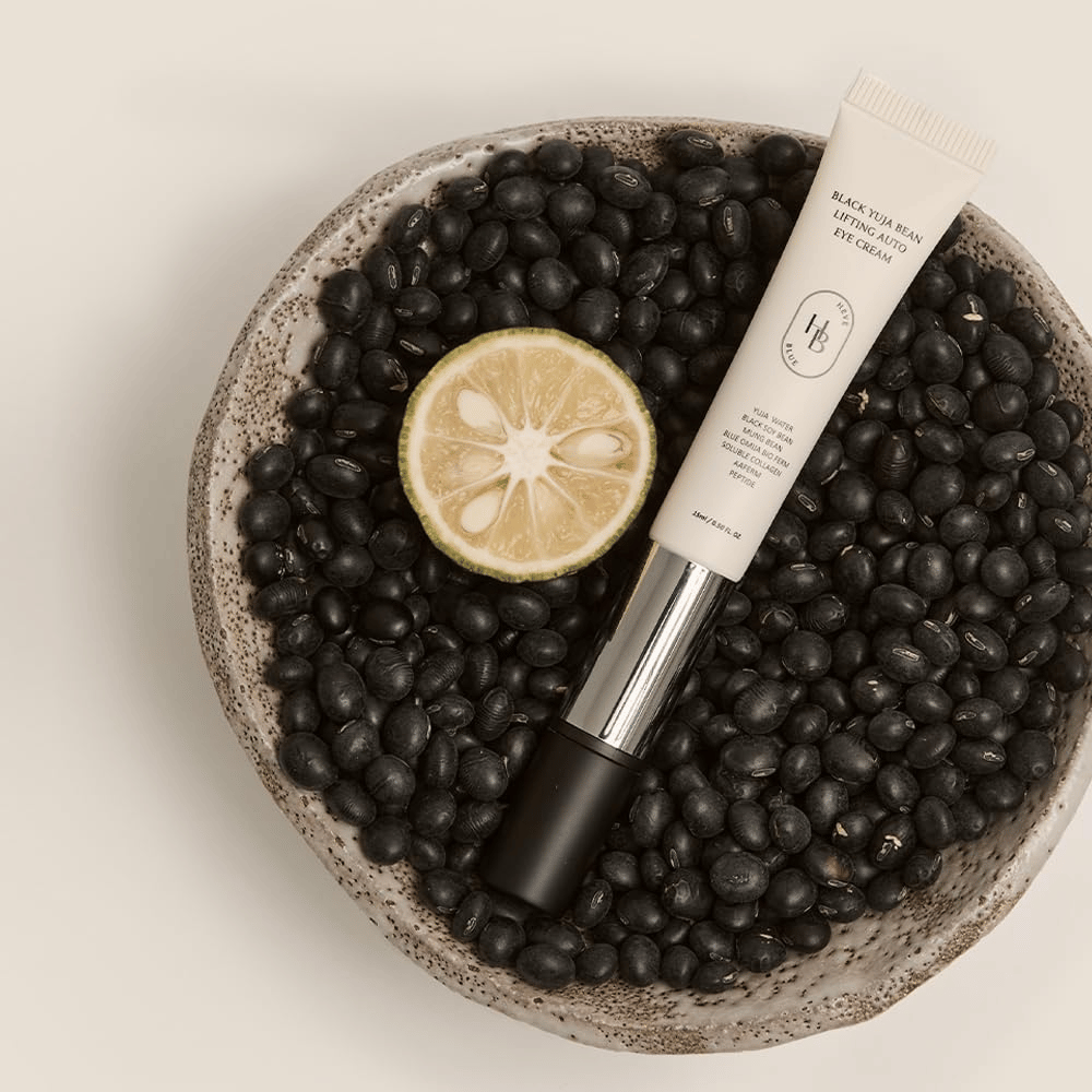 Black Yuja Bean Lifting Auto Eye Cream leži v skledi, napolnjeni s črnim fižolom in prepolovljenim citrusnim sadežem – vizualna predstavitev naravnih sestavin izdelka.